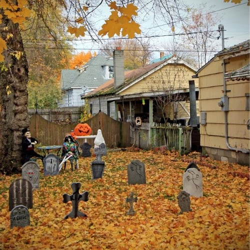 Garten mit Herbstlaub und Gräbern als Halloween-Dekoration. Im Hintergrund ein kleines Haus, während im Vordergrund Grabsteine und eine Kürbislaterne zu sehen sind.