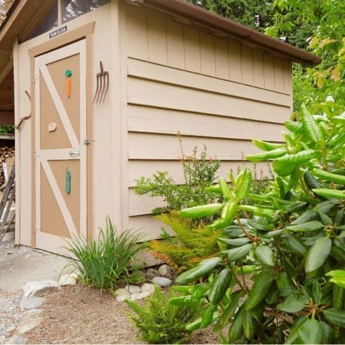 Beiges Gartenhaus mit Holztür, Kiesweg und grünen Sträuchern in einem gepflegten Garten