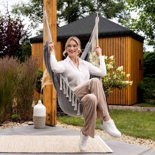 Eine Frau sitzt lächelnd in einer Hängematte von di volio, befestigt an einem Holzbalken. Im Hintergrund ist ein moderner Gartenpavillon aus Holz zu sehen.