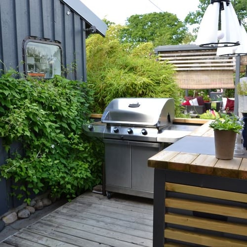 Outdoor-Küche: Eine gemütliche Ecke im Garten mit einem Edelstahl-Gasgrill und einem Holztresen, umgeben von Pflanzen und einer Terrasse aus Holz