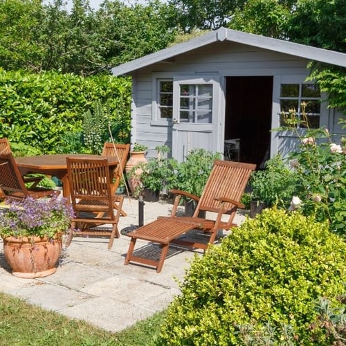 Graues Gartenhaus mit Holzliege, Gartentisch und Stühlen auf einer sonnigen Terrasse mit vielen Pflanzen