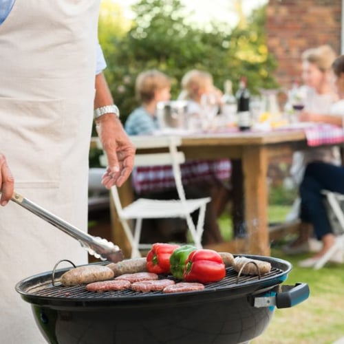 Ein Mann grillt Würstchen und Paprika auf einem runden Grill im Garten auf einem Gartenparty. Im Hintergrund sitzt eine Familie an einem Holztisch.