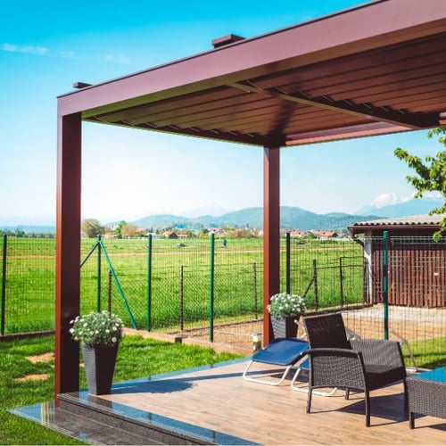 Eine moderne Terrasse mit einem eleganten Pergola, zwei schwarzen Gartenstühlen und einem kleinen Tisch auf einem Holzdeck, mit Blick auf eine grüne Landschaft im Hintergrund.