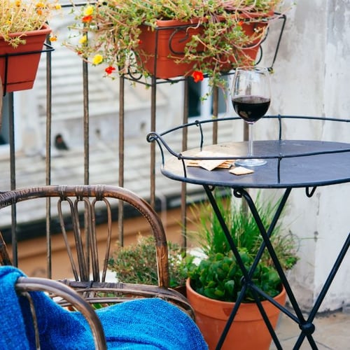 Ein kleiner Balkon mit einem runden Metalltisch und einem Rattansessel. Auf dem Tisch stehen ein Glas Rotwein und ein paar Kekse. Am Geländer hängen Blumentöpfe mit bunten Blumen, und unter dem Tisch stehen Töpfe mit grünen Pflanzen. Eine blaue Decke liegt über dem Sessel.