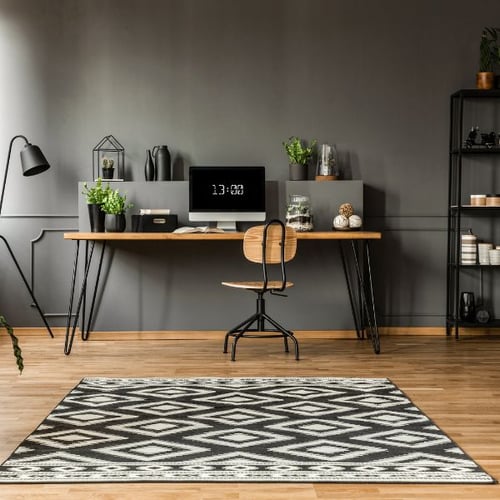 Modern eingerichtetes Homeoffice mit einem großen Holztisch, einem schwarzen Metallstuhl und Dekorationselementen wie Pflanzen und Keramikvasen. Ein schwarz-weißes Teppichmuster liegt auf dem Holzboden, und ein minimalistisches Regal steht an der Wand.