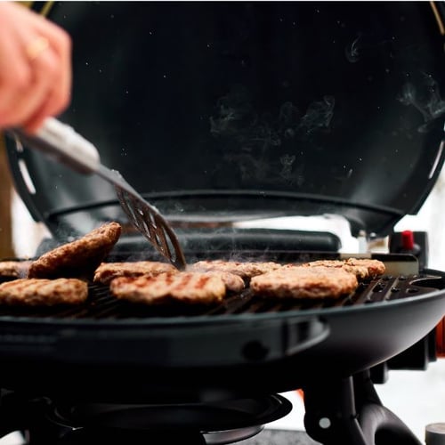 Burger-Patties auf einem geöffneten Gasgrill mit Dampf, eine Hand dreht die Patties mit einem Grillwender um im Winter.