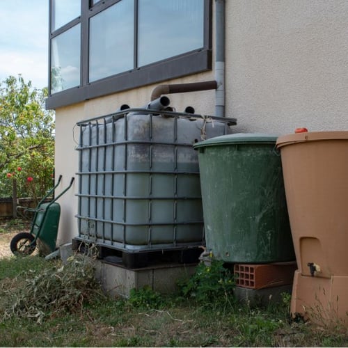 Drei große Wassertanks stehen neben einem Haus. Einer ist ein quadratischer IBC-Tank, die anderen zwei sind runde, grüne und braune Regentonnen.