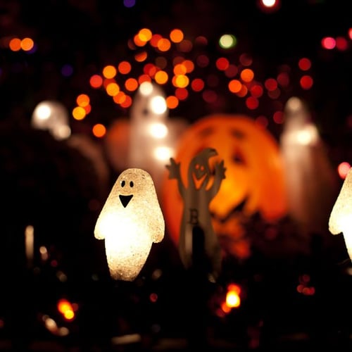 Beleuchtete Halloween-Geisterdekorationen vor einem Hintergrund aus bunten Lichtern. Die Geisterfiguren leuchten im Dunkeln und sind umgeben von orangefarbenen und violetten Lichtern.