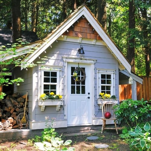 Kleines graues Gartenhaus mit weißen Fenstern, Blumenkästen und dekorativem Türkranz zwischen Bäumen
