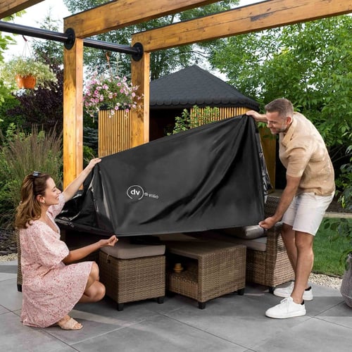 Kobieta i mężczyzna wspólnie zakładają duży, czarny pokrowiec z logo di volio na zestaw mebli z technorattanu. Zdjęcie prezentuje praktyczny sposób na to, jak zabezpieczyć meble ogrodowe przed wiatrem, deszczem i kurzem na tarasie pod drewnianą pergolą.