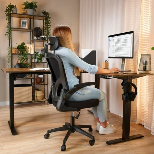 Kobieta pracująca przy biurku w ergonomicznym fotelu z zagłówkiem i podłokietnikami, w jasnym domowym biurze z roślinami – inspiracja dla tych, którzy zastanawiają się, jaki fotel ergonomiczny do 1000 zł wybrać do home office.