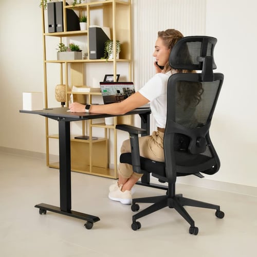 Kobieta siedzi przy biurku na ergonomicznym fotelu z wysokim oparciem i regulowanym zagłówkiem, pracując przy laptopie. Doskonała ilustracja, jaki fotel ergonomiczny do komputera zapewnia wygodę i prawidłową postawę.